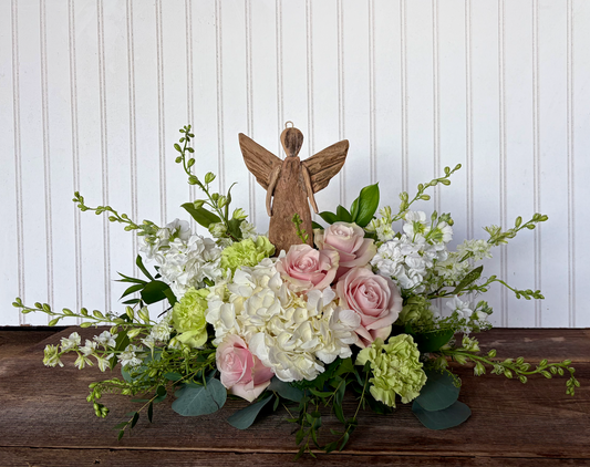 A rustic floral centerpiece featuring pink and white roses, hydrangeas, green foliage, and a small wooden or glass angel figurine.