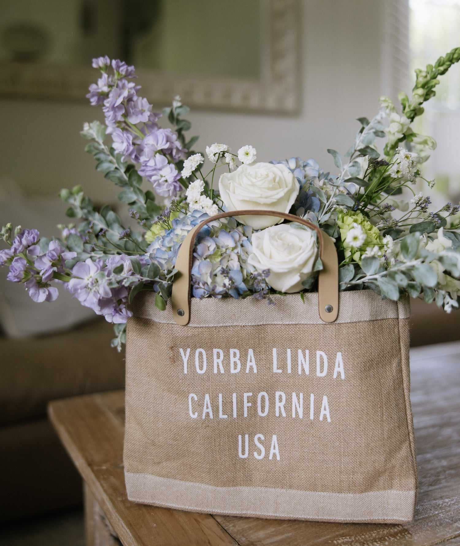A natural burlap tote bag with 'YORBA LINDA CALIFORNIA USA' printed in white, filled with white roses, lavender, and green foliage, sitting on a wooden table in a softly lit room.
